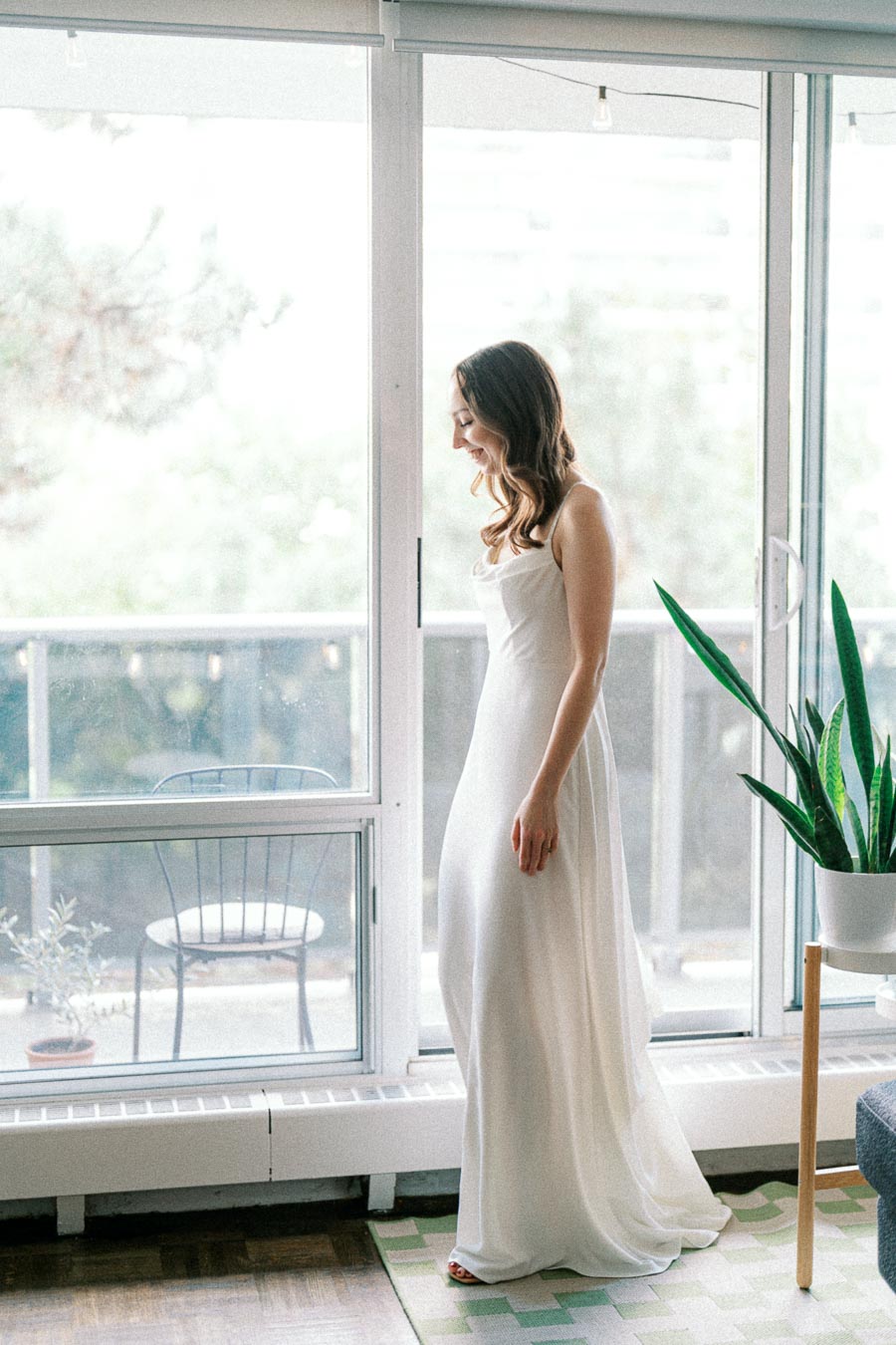 A woman in a long white dress stands thoughtfully in a sunlit room next to large glass doors, with a potted plant and outdoor chair visible on a balcony in the background.