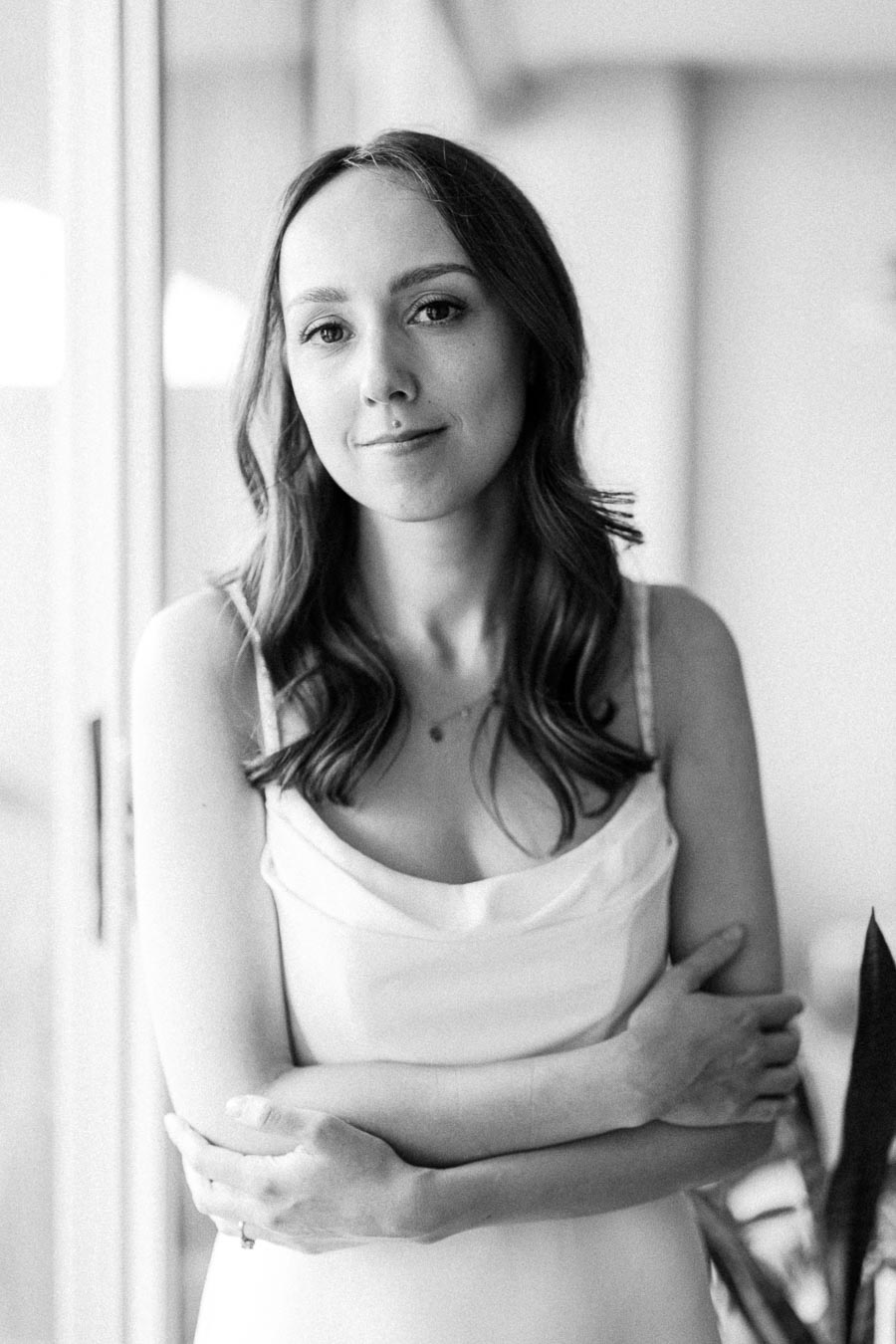 A black and white portrait of a woman with long hair, wearing a sleeveless dress, standing with folded arms in front of a window, exuding a calm and confident demeanor.