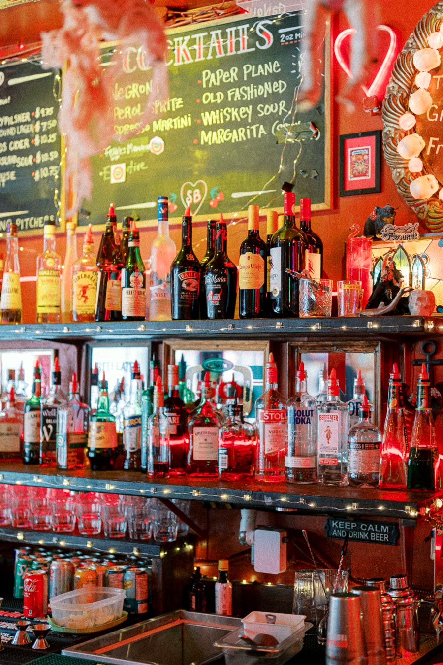A vibrant bar interior showcasing a variety of liquor bottles, with a chalkboard menu displaying cocktail options like Negroni and Margarita, illuminated by soft red lighting for a cozy atmosphere.