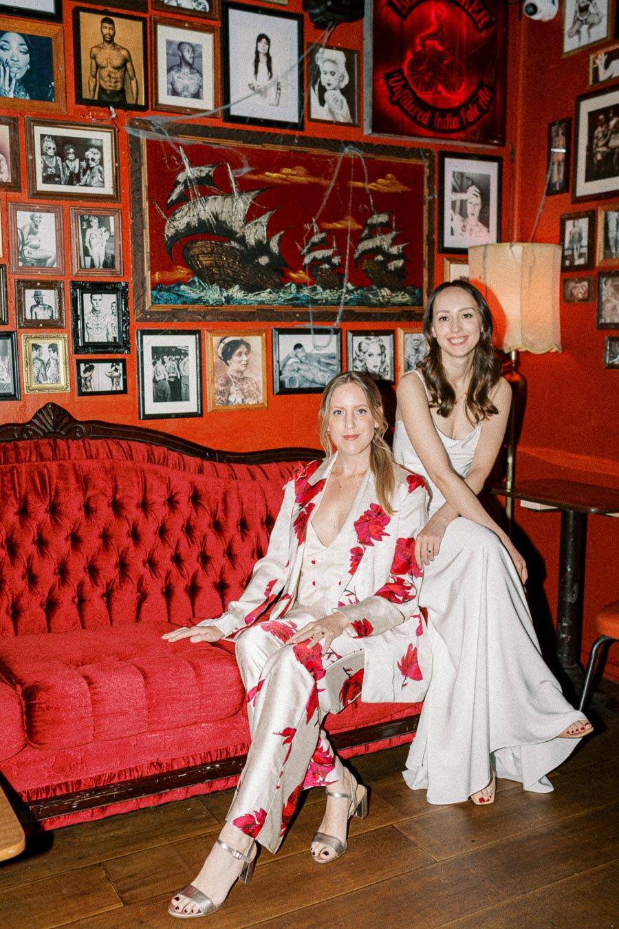 Two women in elegant outfits sit on a vintage red velvet couch in a room with vibrant orange walls adorned with numerous framed portraits and ship artwork, creating a stylish and eclectic atmosphere.