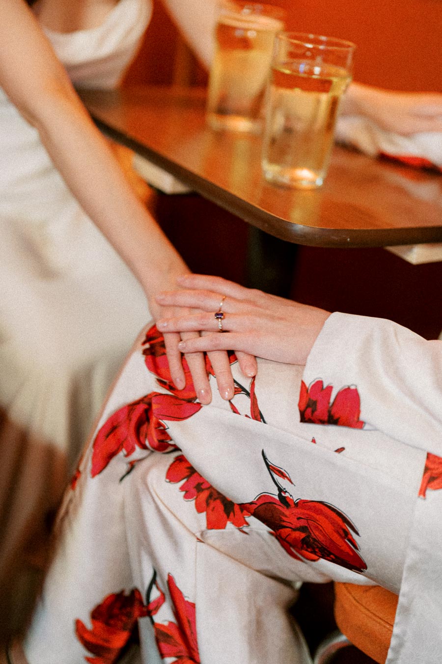 Intimate close-up of two people holding hands across a table, one wearing a floral-patterned dress and a ring, with two glasses of drink in the background, conveying connection and warmth.
