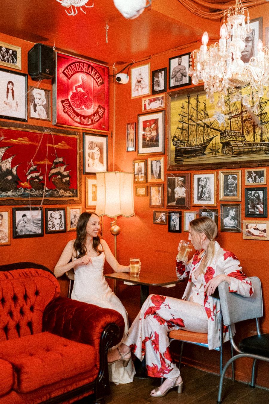 Two women enjoying drinks in a vintage-themed bar with orange walls adorned with eclectic artwork, including a neon sign and framed pictures. One woman is in a white dress, and the other is in a floral suit, seated in a cozy room with a chandelier and plush red couch.