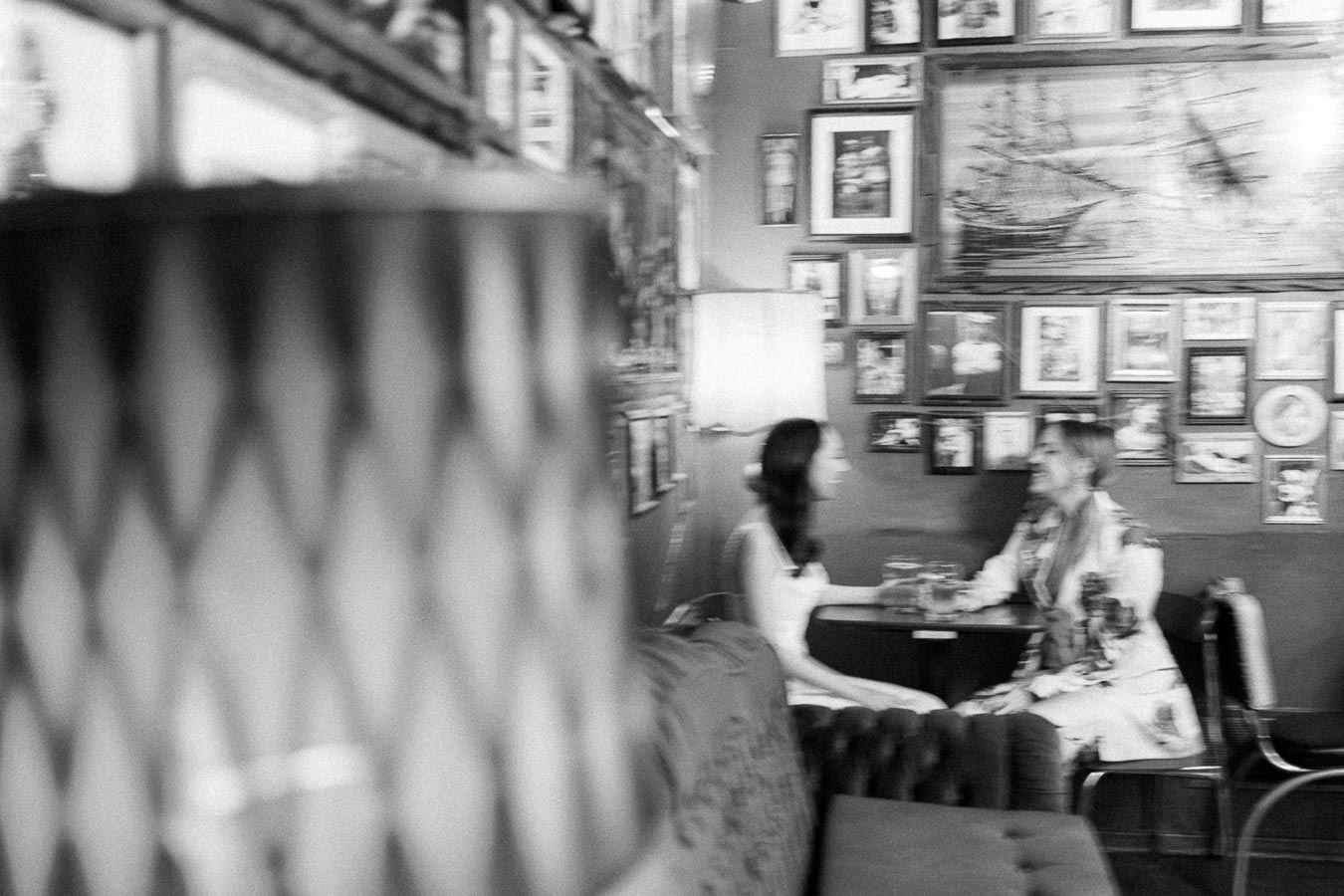 Blurry black and white image of two women sitting at a table in a cozy café, surrounded by framed artwork on the walls.