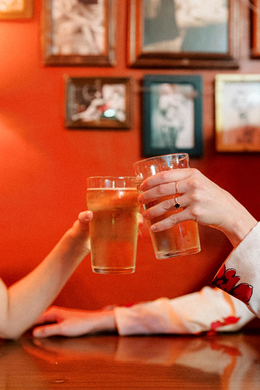 Two people clinking glasses of beer in a cozy, warmly lit pub with vintage photos on a red wall, celebrating a moment together.
