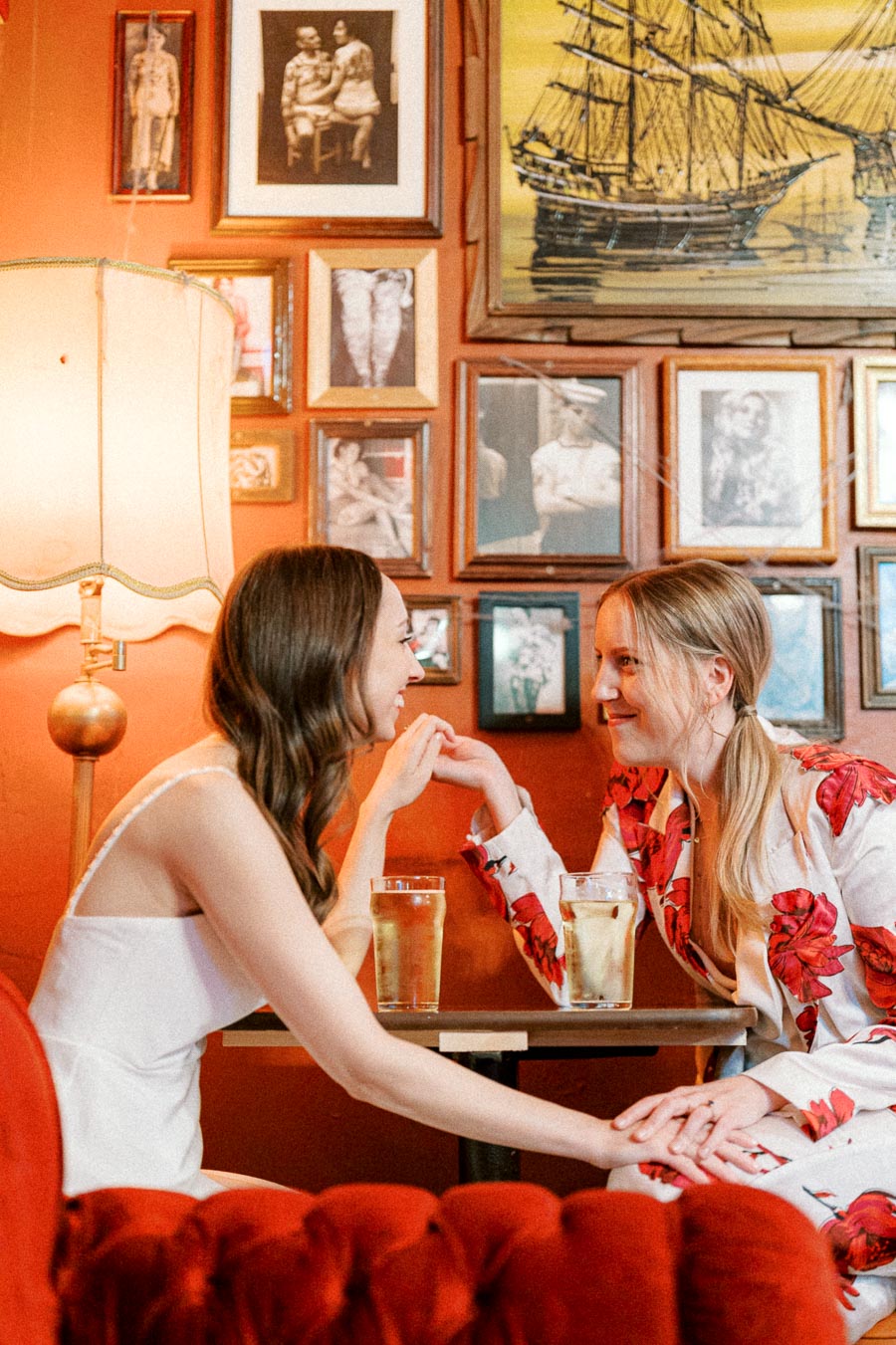 Two women smiling and holding hands at a cafe table with drinks, surrounded by vintage decor and framed pictures.