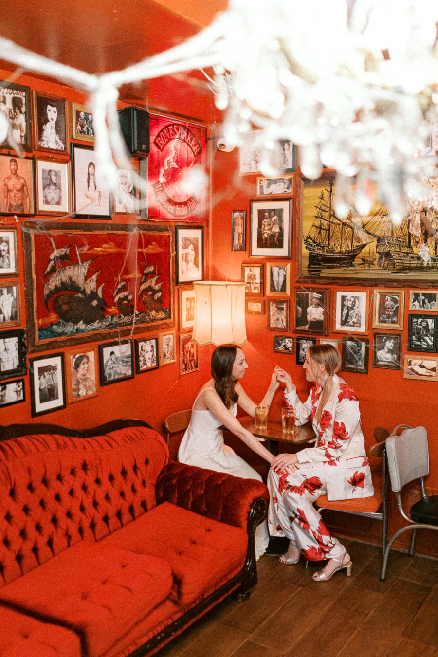 Two women enjoying drinks in a cozy, vintage-themed bar with red decor and eclectic wall art.