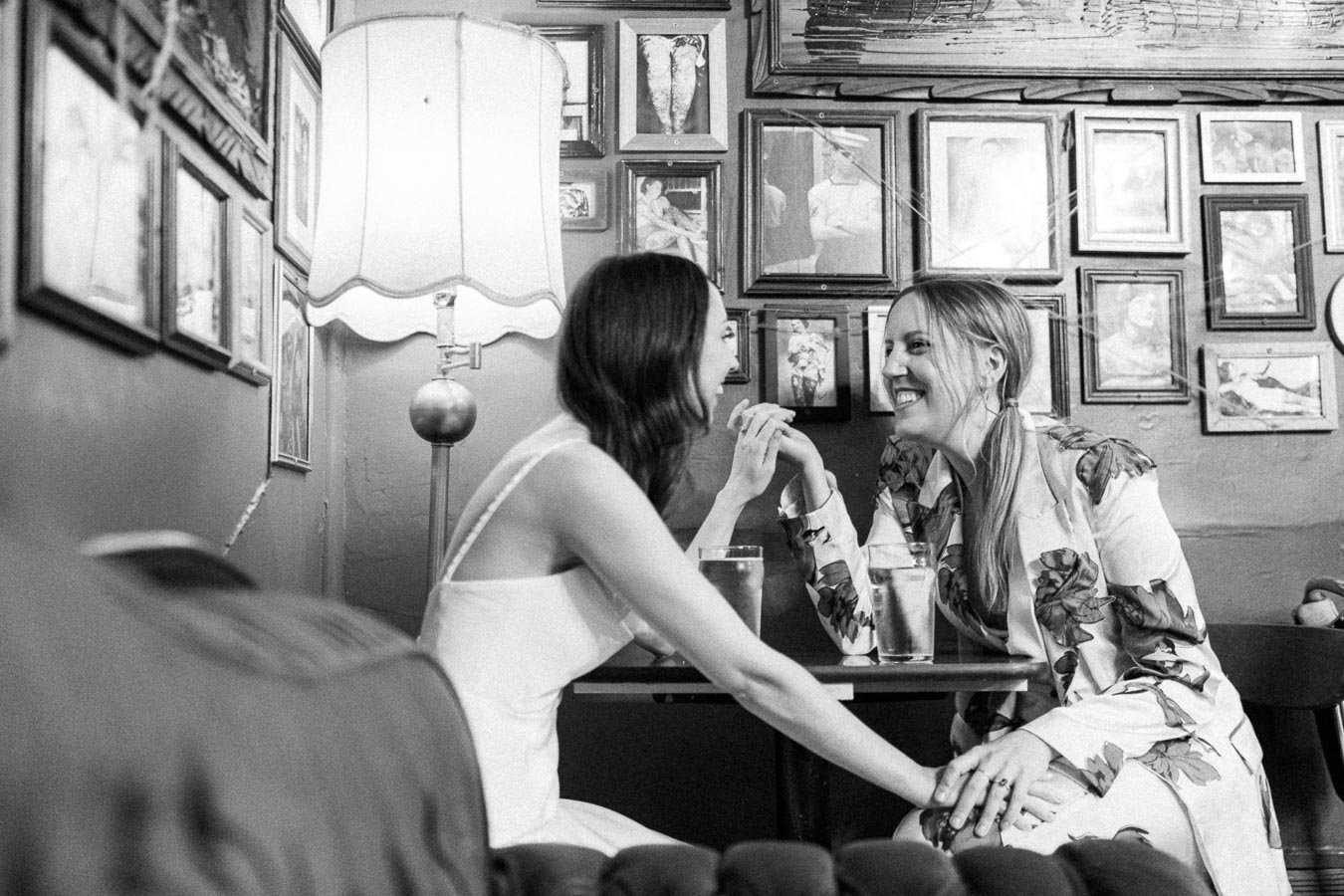 Two women sitting at a cozy cafe table, laughing and holding hands, surrounded by framed art and a vintage lamp, creating a warm and intimate atmosphere.