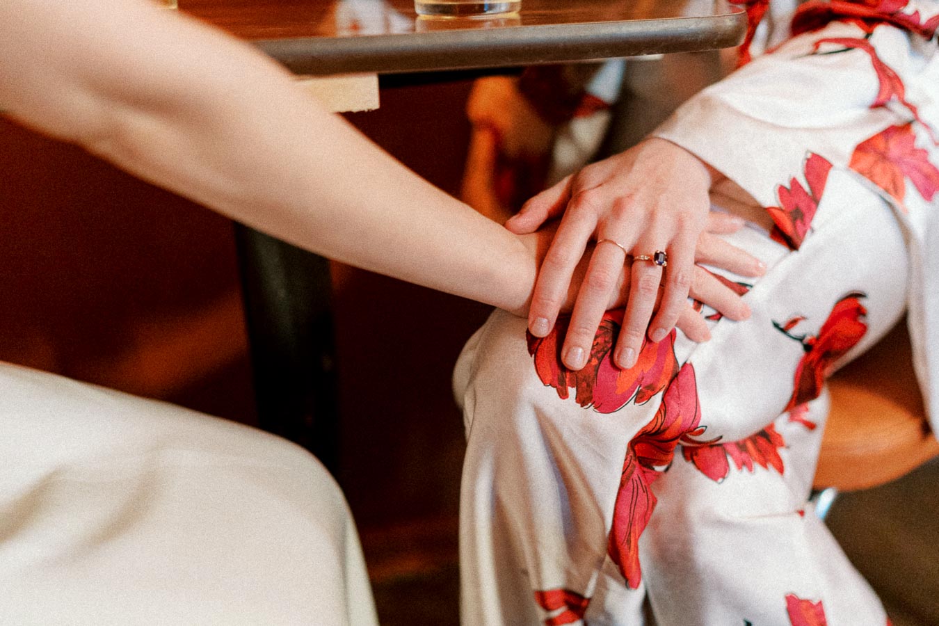 Two people seated at a table, one wearing floral-patterned clothing with a ring on their finger, gently holding hands, conveying a sense of connection and intimacy.