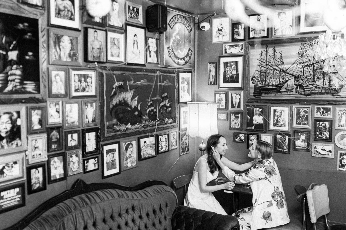 Black and white image of two women sitting in an eclectic room filled with framed photographs and artwork. They appear engaged in a warm, joyful conversation, surrounded by vintage decor including a tufted sofa and a standing lamp. The walls are adorned with diverse portraits and paintings, creating an intimate and nostalgic atmosphere.