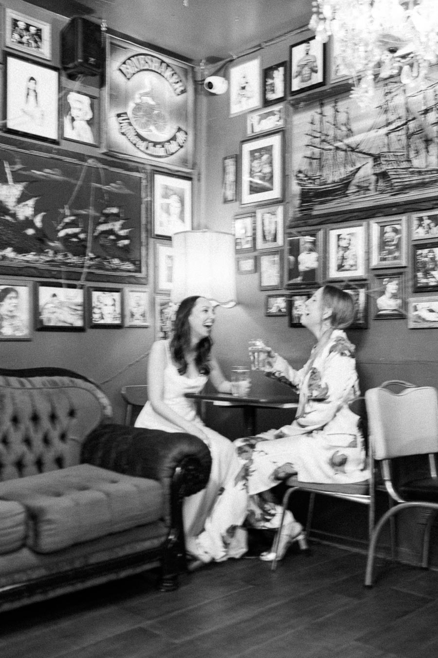 Two women laughing and enjoying drinks in a cozy, art-filled room with a vintage ambiance. The walls are adorned with an eclectic mix of framed photographs and paintings, while the women are seated on comfortable furniture, creating a relaxed and joyful atmosphere.