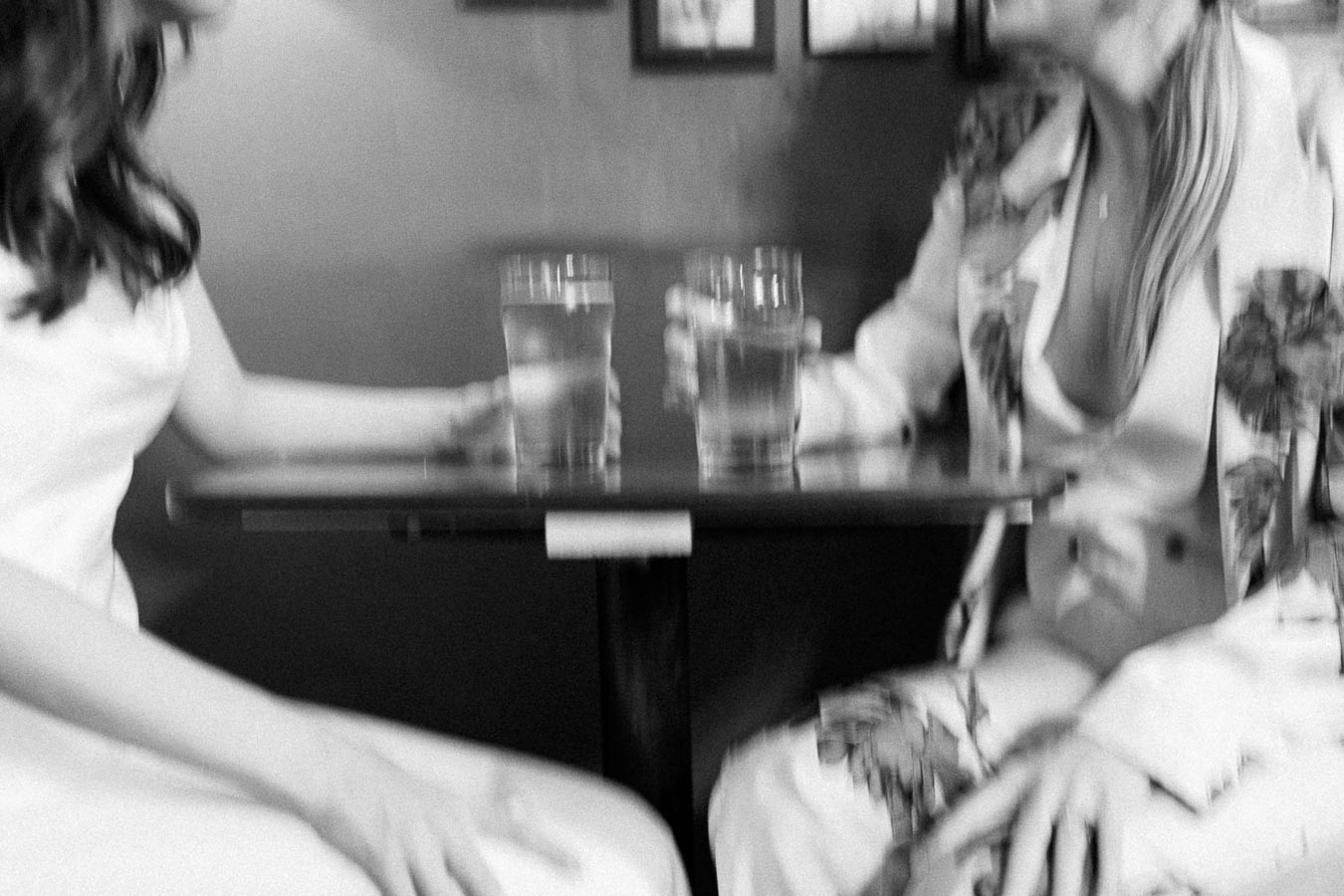 Two women sitting at a table in a cafe, holding glasses of water. The image is in black and white, capturing a candid and relaxed moment.