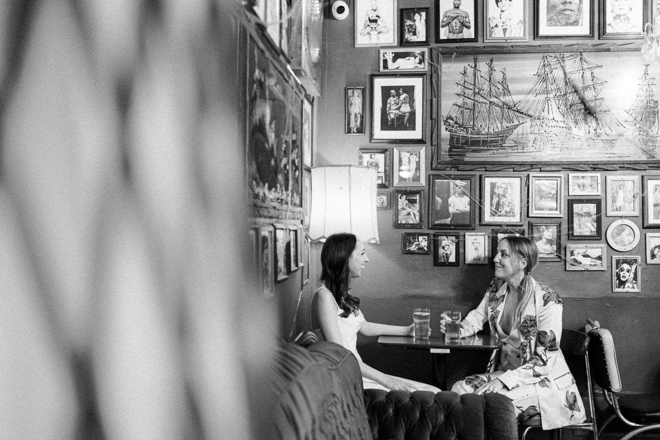 Black and white image of two women sitting at a small table in a cozy cafe, surrounded by eclectic wall art and framed pictures, engaged in conversation and enjoying drinks.
