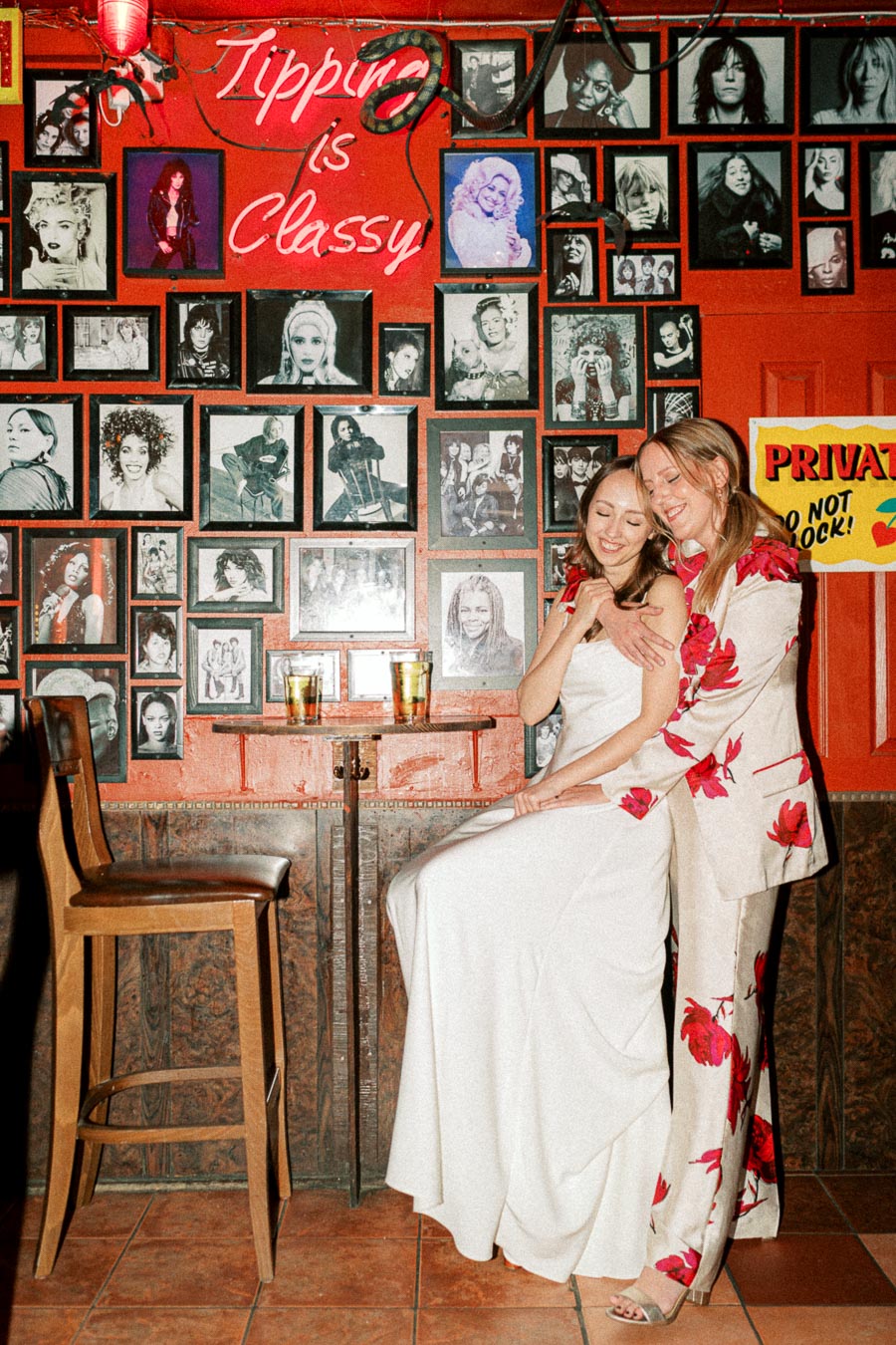Two women in elegant outfits smiling in a bar with a wall of celebrity photos. A neon sign reads Tipping is Classy, and two drinks on a small table enhance the cozy atmosphere.