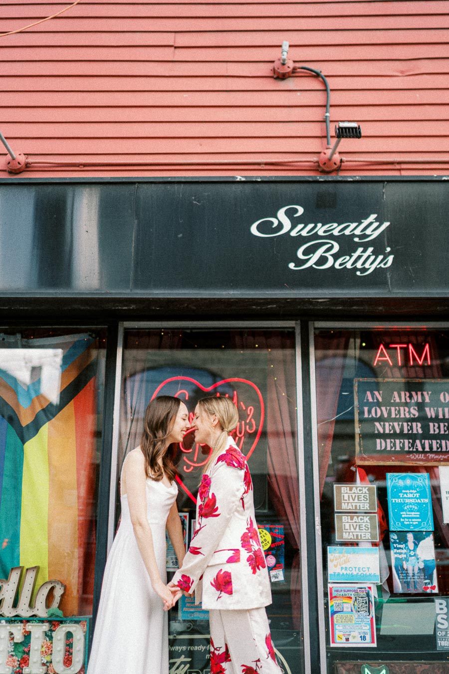 A couple stands close together, holding hands and laughing in front of a storefront with a colorful Pride flag and various posters, under the sign that reads Sweaty Betty's.