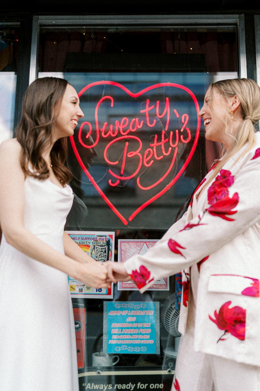 Two women smiling and holding hands in front of a neon sign that reads Sweaty Betty's, with vibrant clothing and a heart design in the background.