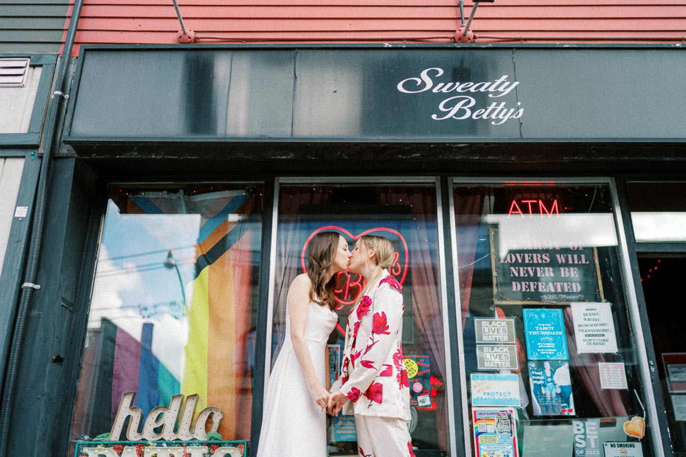 Two people share a kiss in front of Sweaty Betty's storefront adorned with colorful pride flags and heart-shaped neon sign, capturing a moment of love and diversity.