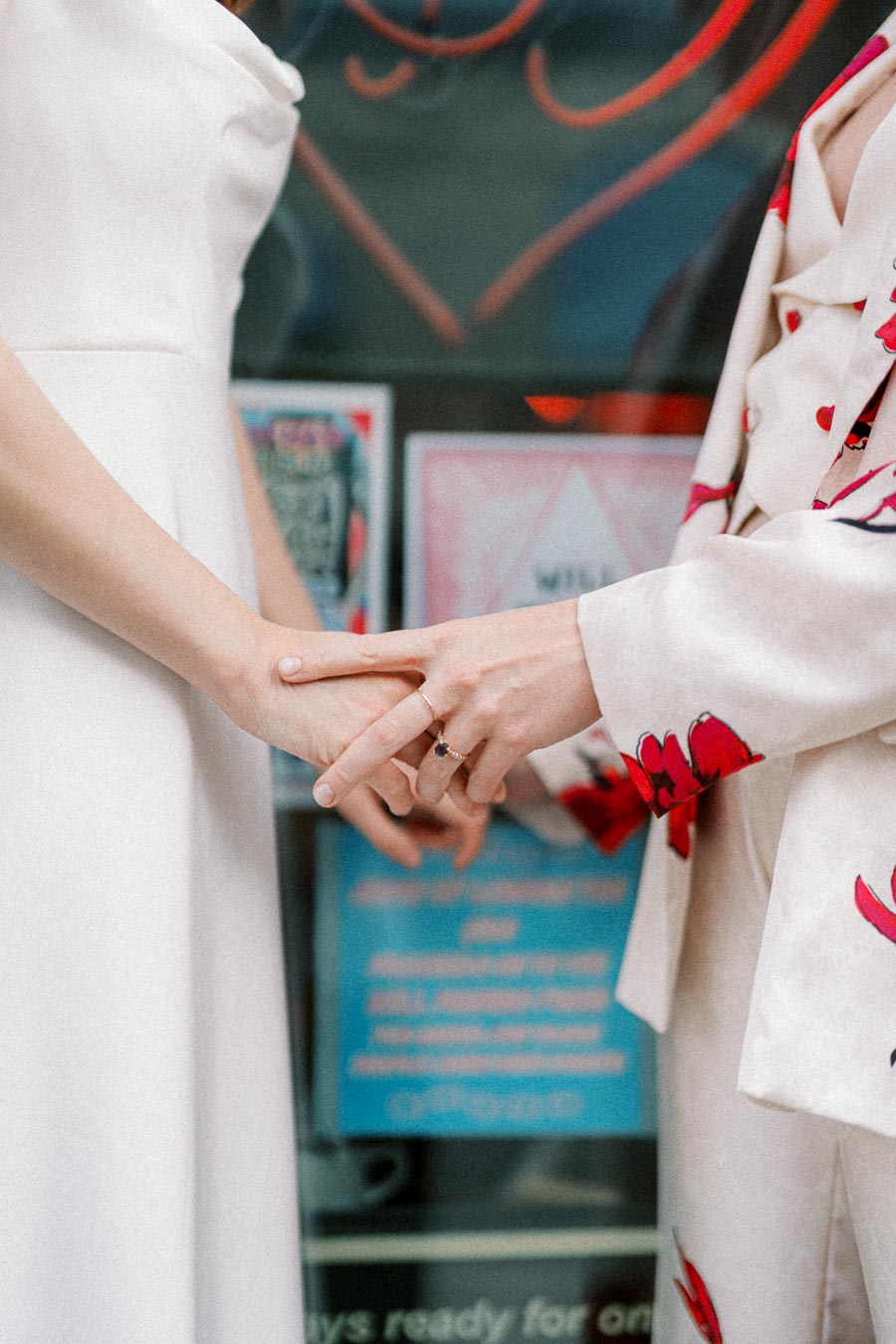 A close-up of two people holding hands, symbolizing love and commitment. One person is wearing a wedding dress, while the other wears a floral suit, both displaying a bond against a colorful backdrop.