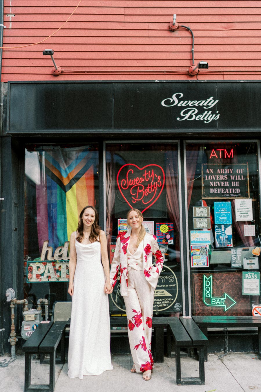 Two individuals stand hand in hand outside Sweaty Betty's, a venue with colorful signage and LGBTQ+ pride decorations. One wears a white dress, the other a floral suit, showcasing a stylish and inclusive atmosphere.