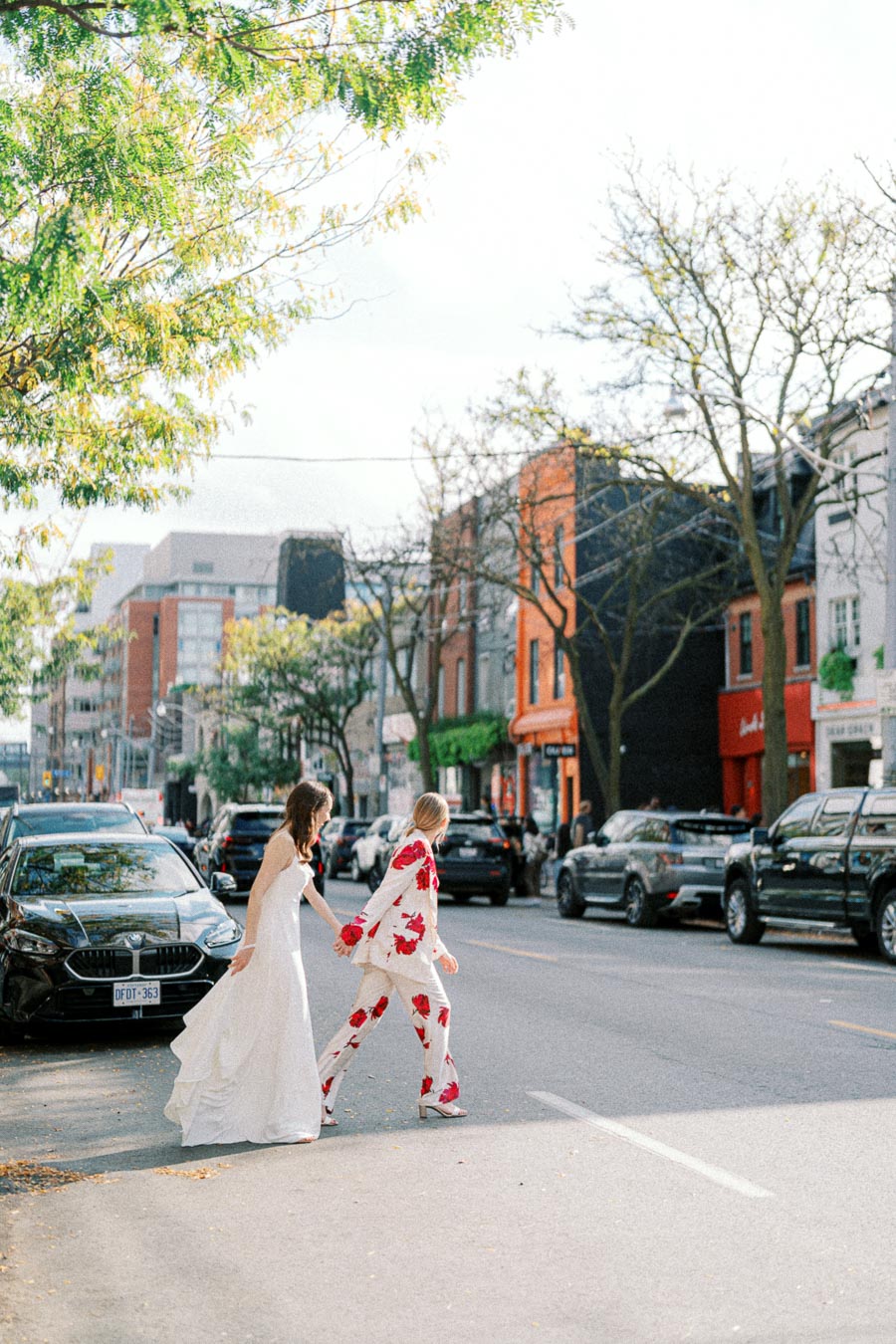 Wedding couple crossing a city street, with the bride in a white dress and the partner in a floral suit, surrounded by urban buildings and parked cars, under a sunny sky with green trees.