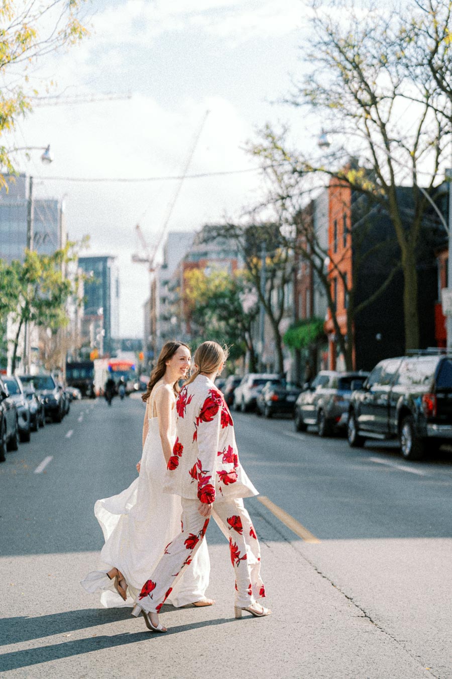 Two women walking down a city street, one in a flowing white dress and the other in a floral-patterned suit, surrounded by parked cars and urban buildings on a sunny day.