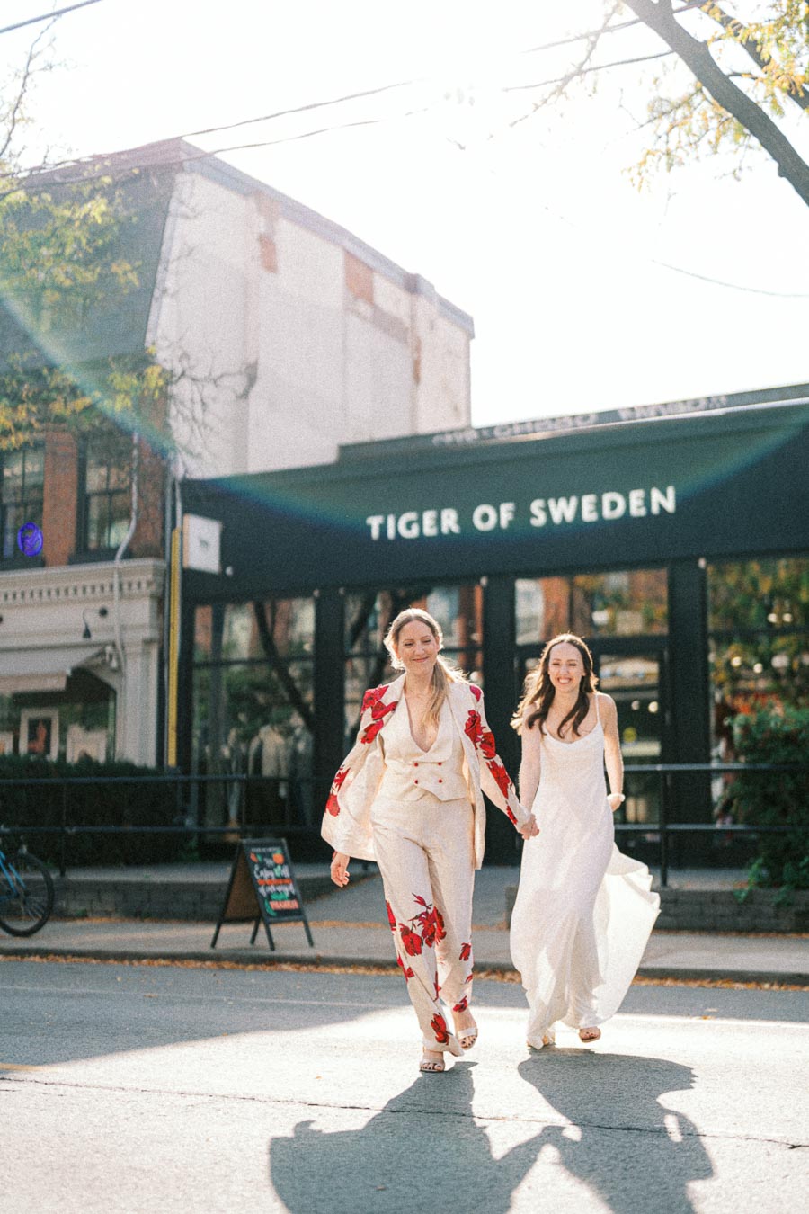 Two people in elegant wedding attire joyfully walking hand in hand on a sunny day in front of the Tiger of Sweden store, with soft light and shadows enhancing the celebratory mood.
