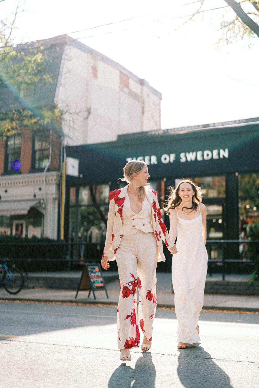 Two women in stylish outfits walking hand in hand down a city street, with a Tiger of Sweden storefront in the background. The sunlight creates a warm and bright atmosphere, enhancing the joyful moment.