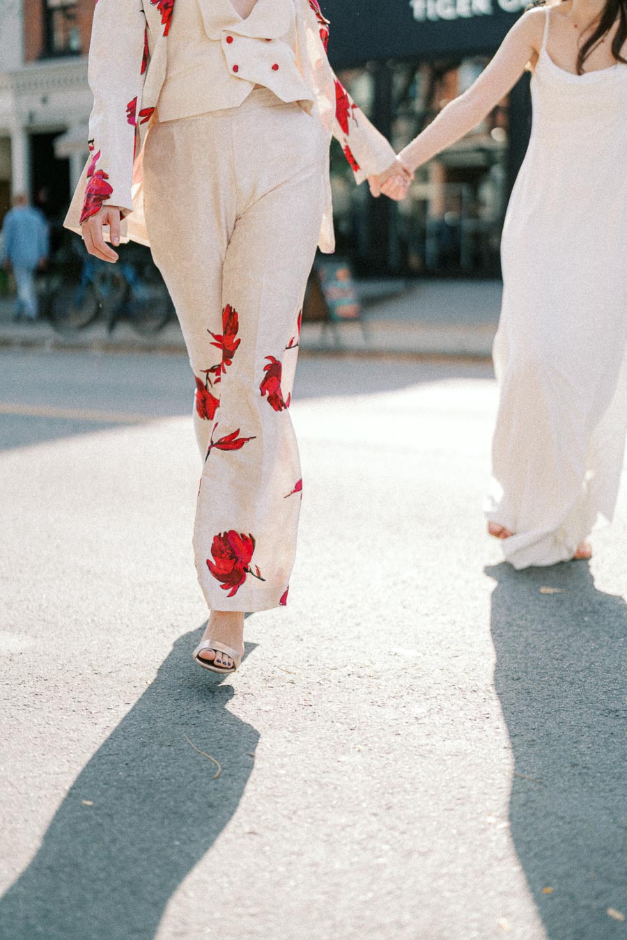 Stylish woman in floral suit walking outdoors in sunlight, holding hands with another woman in a white dress.