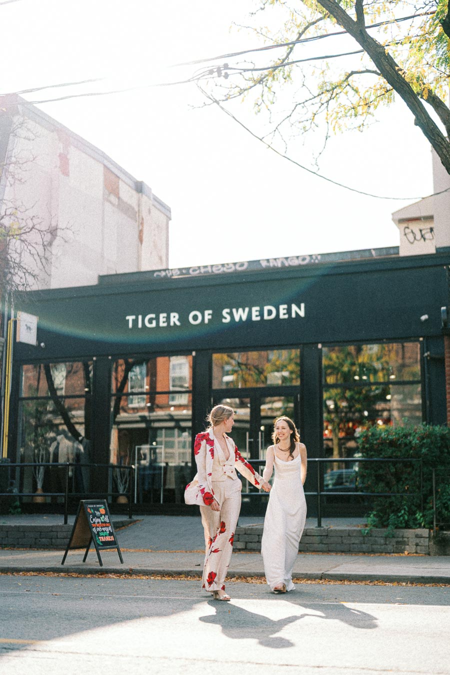 Two people walking and smiling outside the Tiger of Sweden store, showcasing stylish outfits in a sunny urban setting.