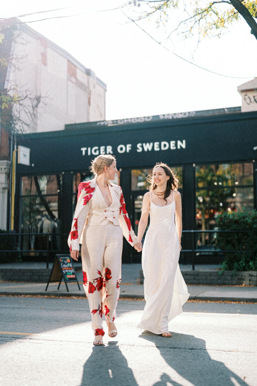 Two women in stylish outfits holding hands and smiling while walking on a sunny street in front of a Tiger of Sweden store.