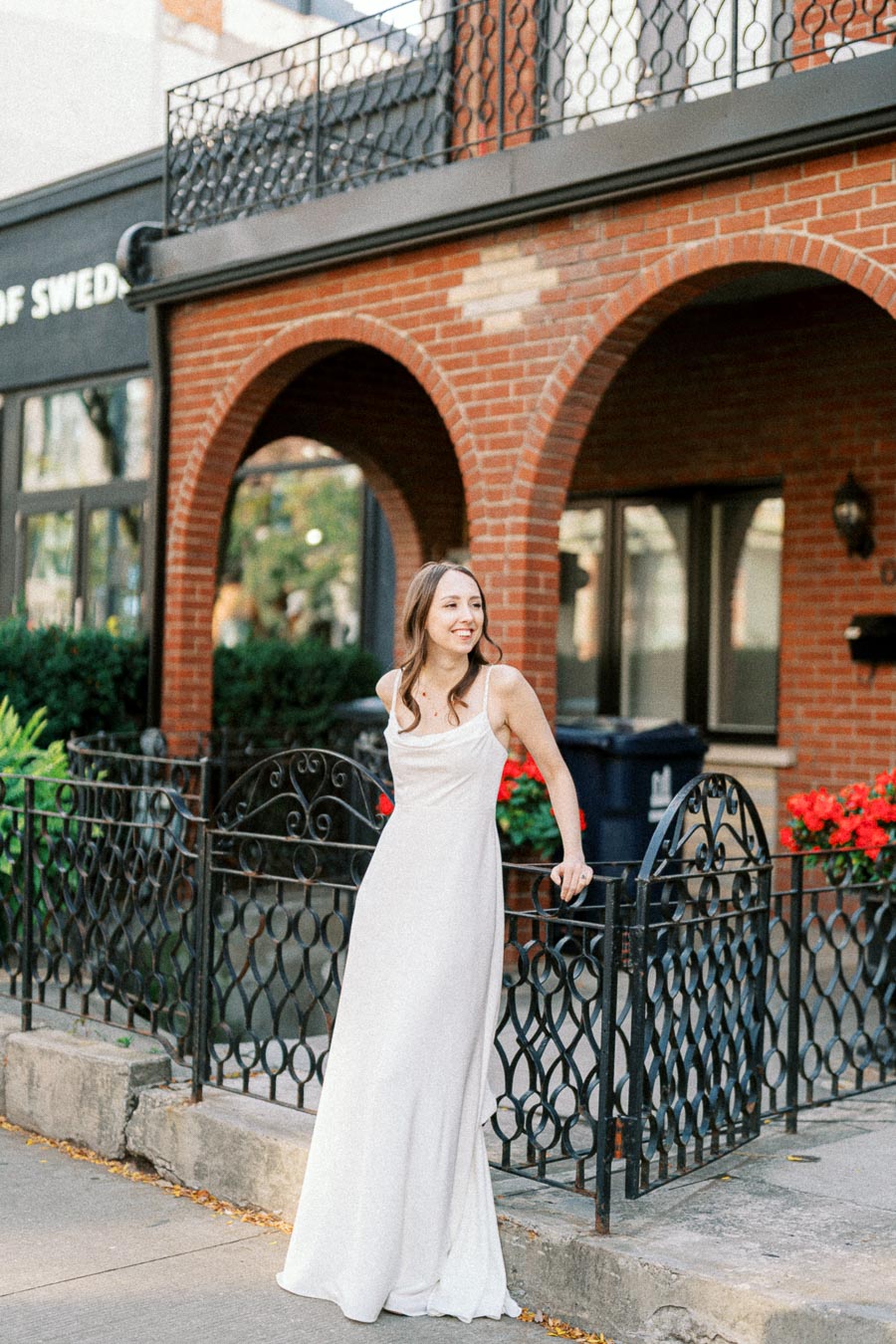 Young woman in a white dress smiling while leaning on a decorative wrought iron fence, with a brick building and vibrant flowers in the background.