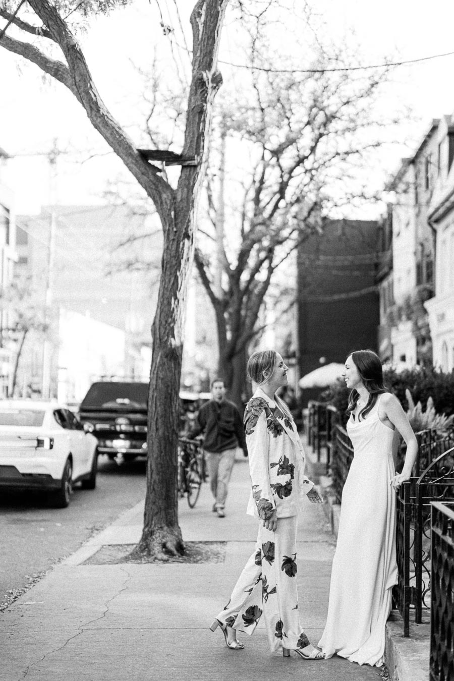 Black and white image of two women in elegant attire having a conversation on a city sidewalk, surrounded by parked cars and trees, capturing an urban street fashion vibe.
