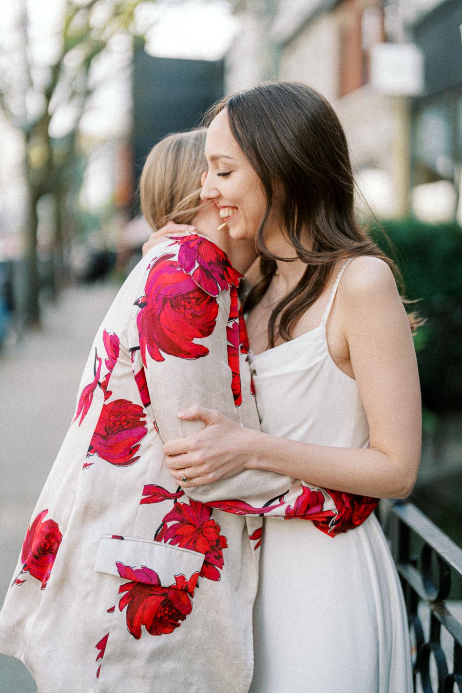 Two women embracing on a city street, one wearing a floral-patterned jacket and the other in a white dress, conveying warmth and friendship.