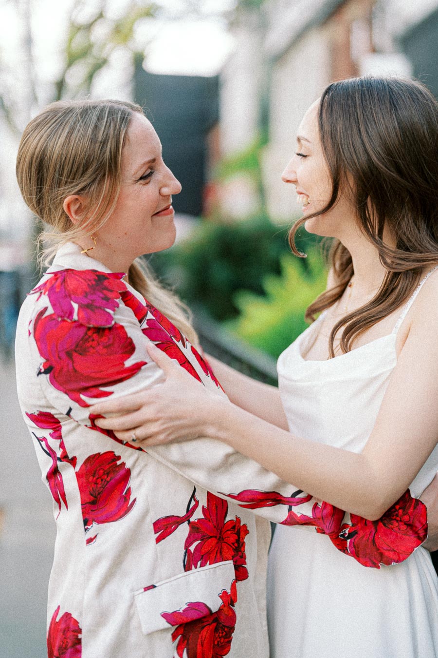 Two women smiling at each other outdoors, one wearing a floral-patterned jacket and the other in a white dress, demonstrating a moment of friendship and joy.