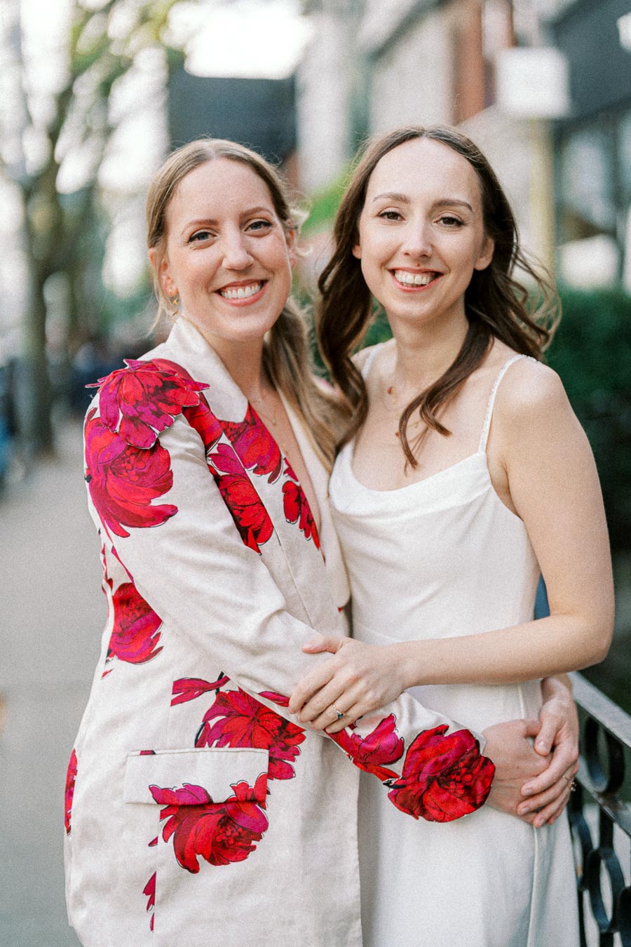 Two women smiling and embracing, one wearing a white dress and the other a floral blazer, standing on an outdoor urban sidewalk.
