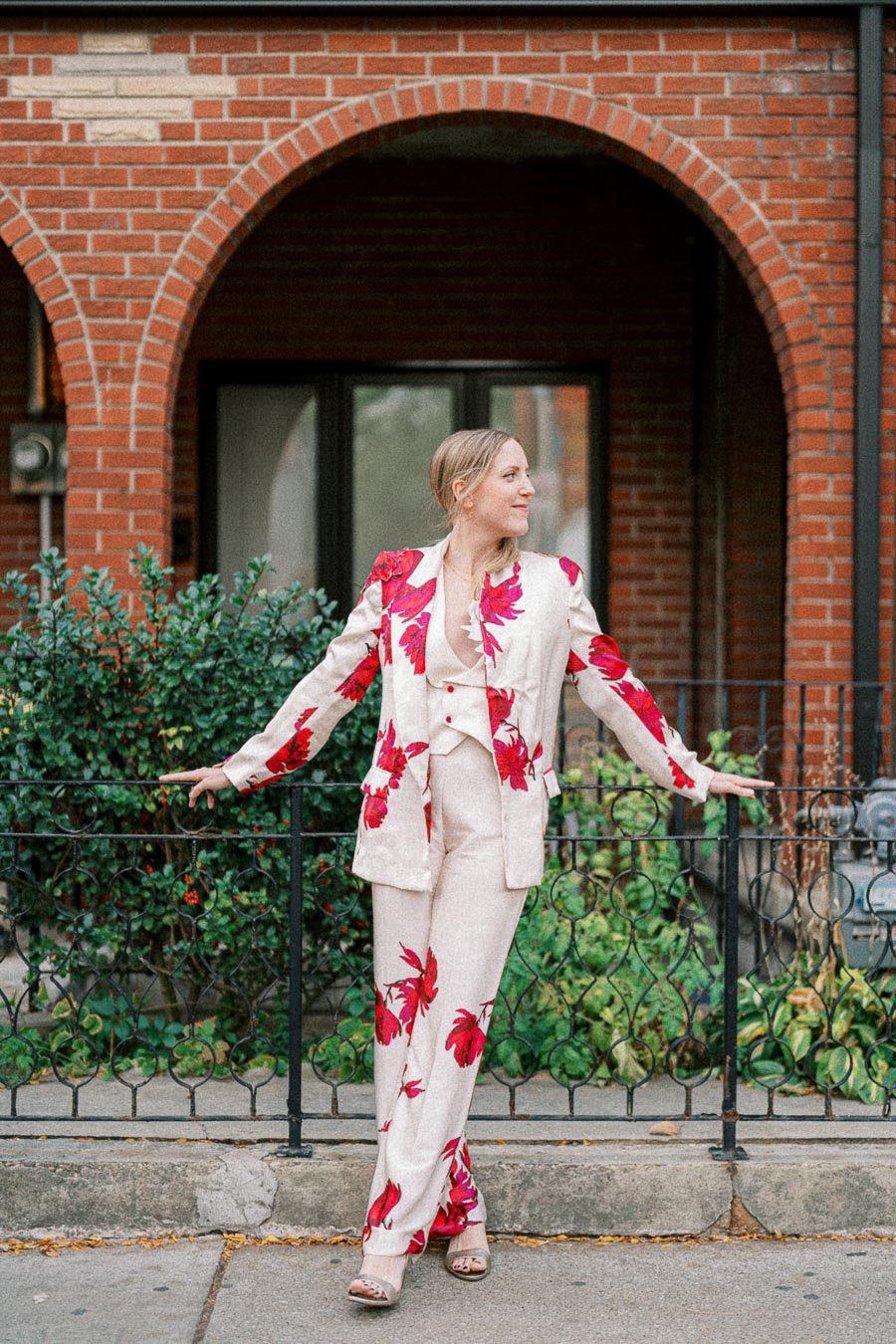 A woman in a stylish cream suit with bold red floral patterns poses confidently in front of a brick building, with lush greenery in the background.
