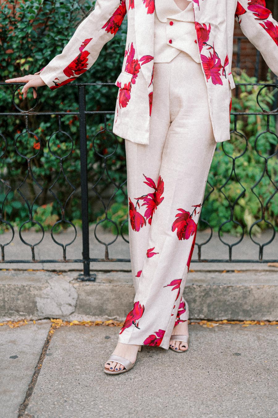 Woman in a stylish beige suit with vibrant red floral patterns, posing against a decorative metal railing, showcasing elegant fashion perfect for a chic urban setting.