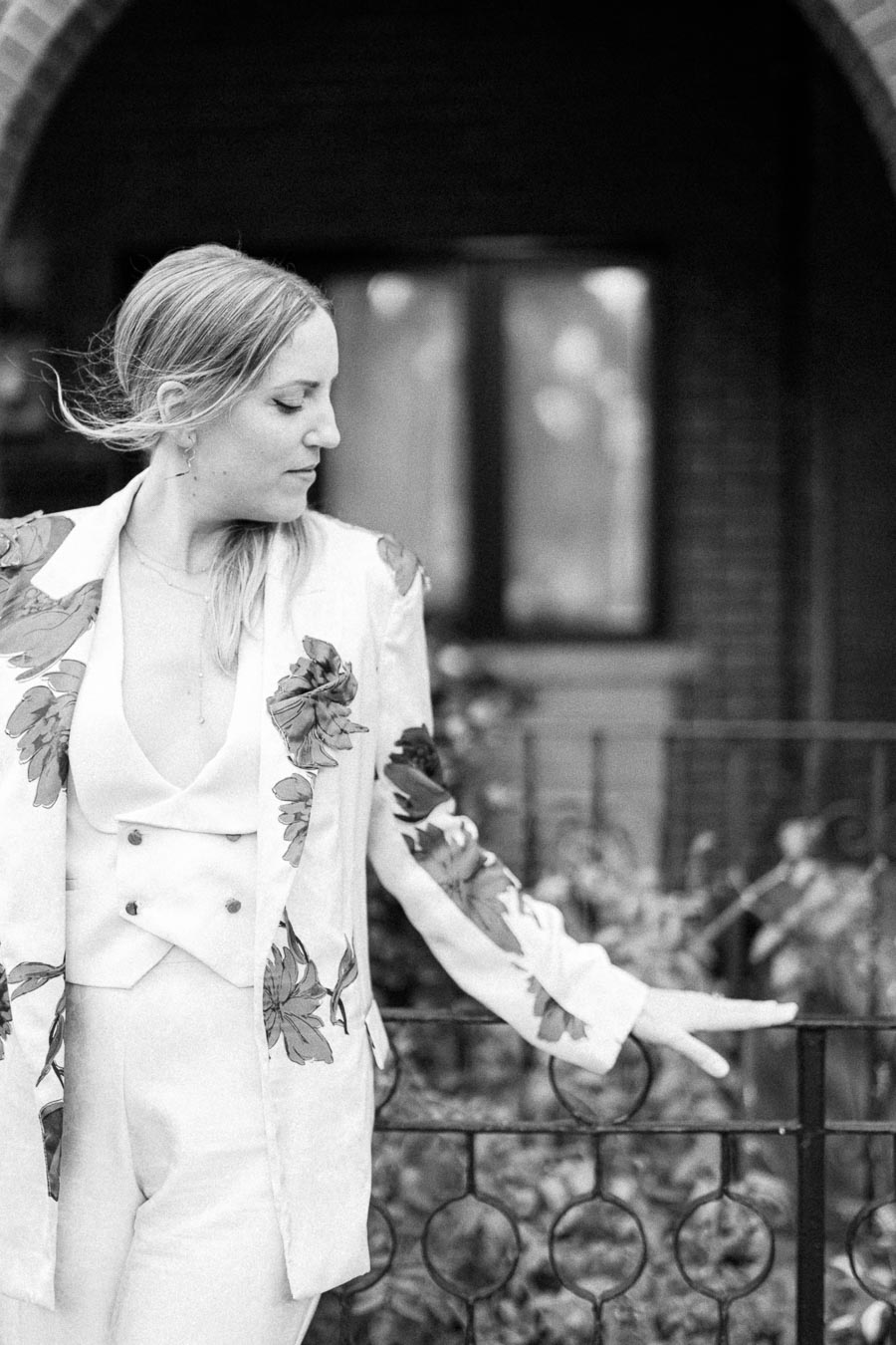 Black and white photo of a woman in a floral-patterned suit, standing outdoors near an iron fence, with a contemplative expression.