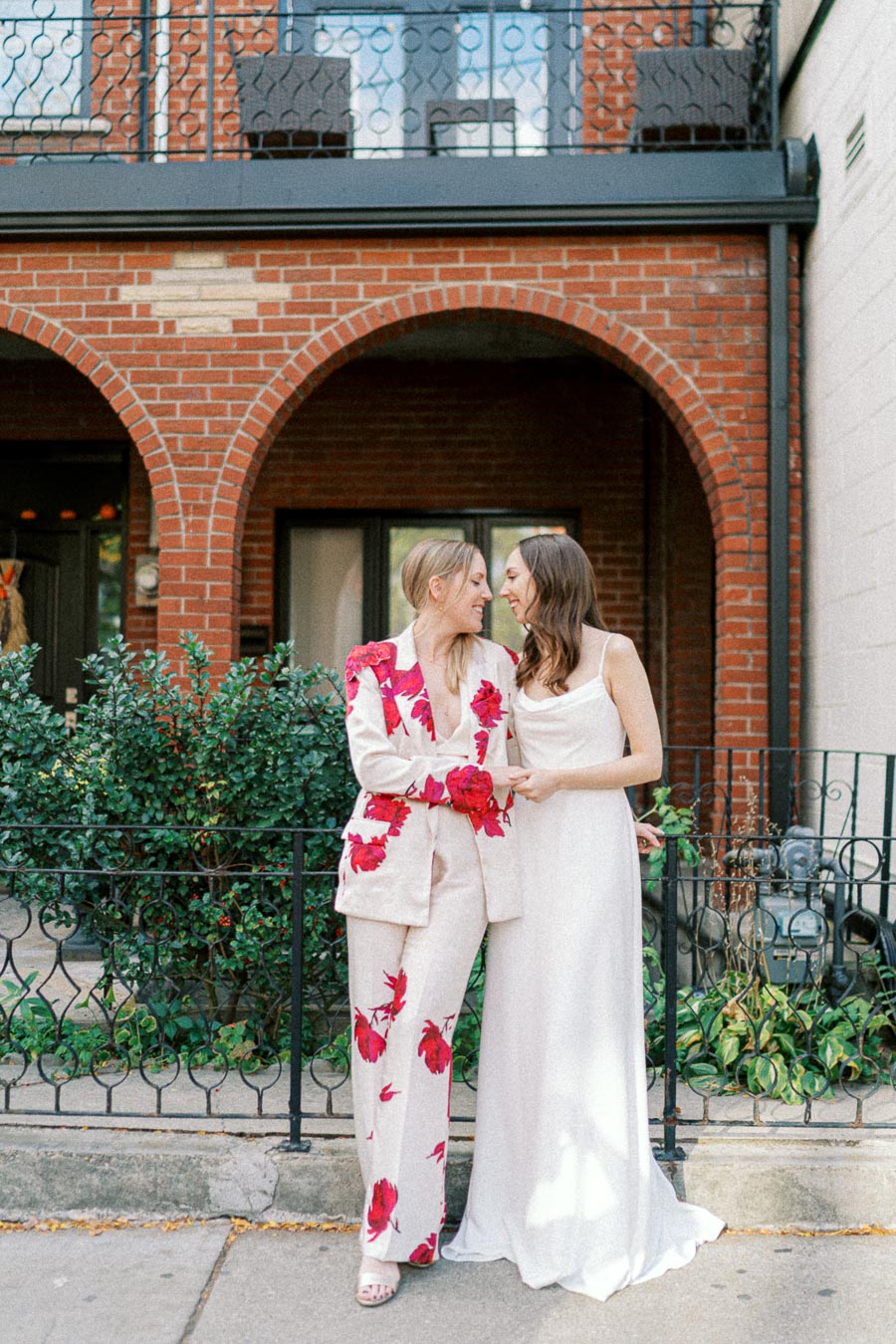 Two women lovingly embrace in front of a brick building, one wearing a floral suit and the other in a white dress, representing a joyful moment and celebration of love.