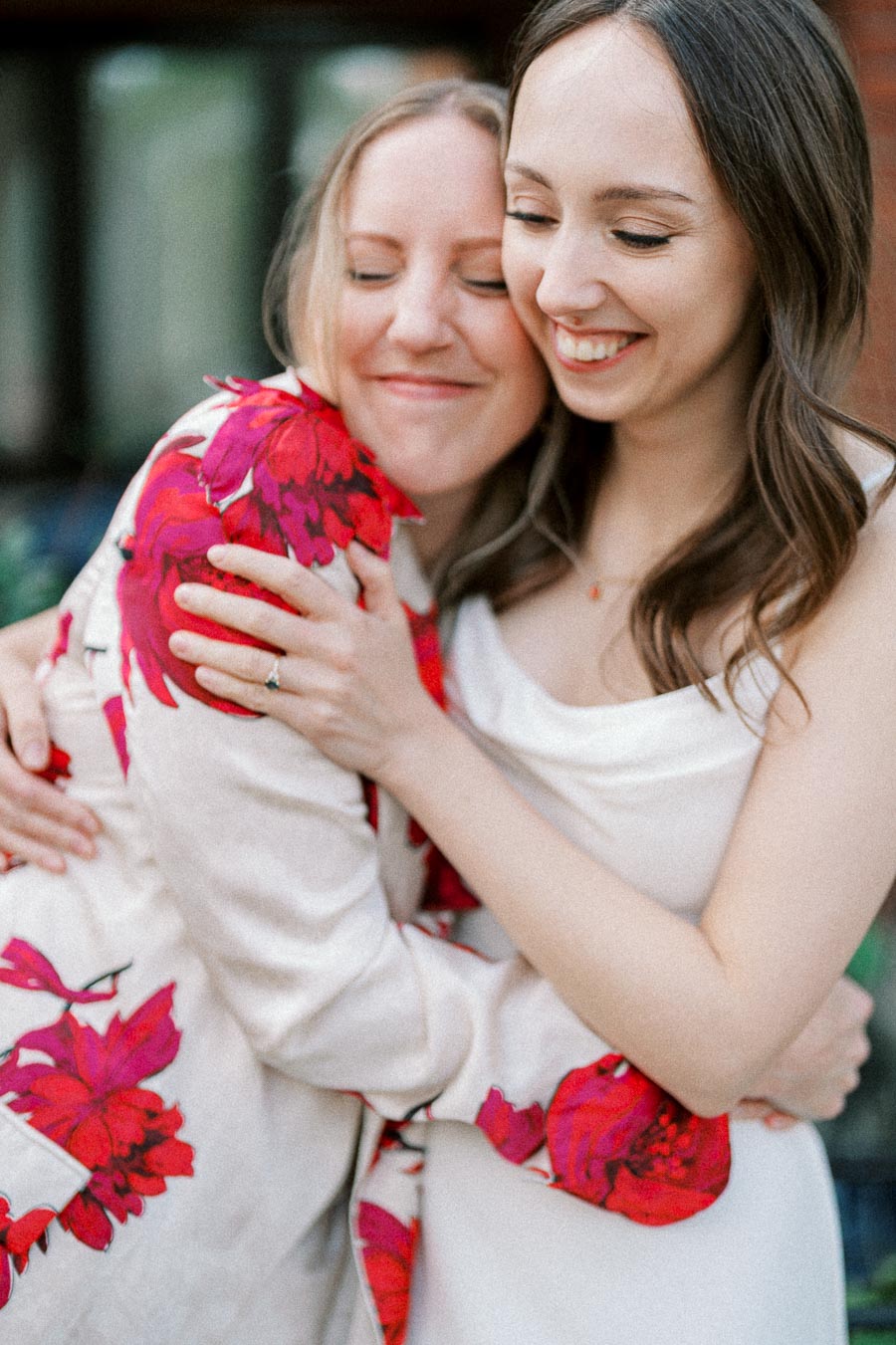 Two smiling women embracing warmly, with one wearing a white dress and the other in a floral jacket.