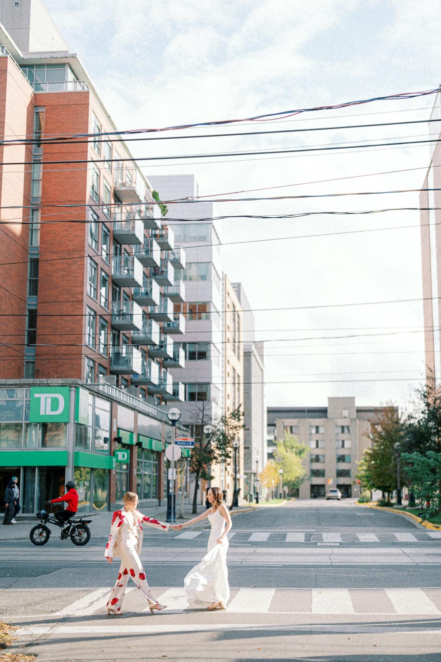 Two women wearing elegant outfits dance in the middle of a sunny urban street. Surrounding them are modern apartment buildings and a bank. A person rides a motorbike in the background. The scene captures a vibrant city life moment.