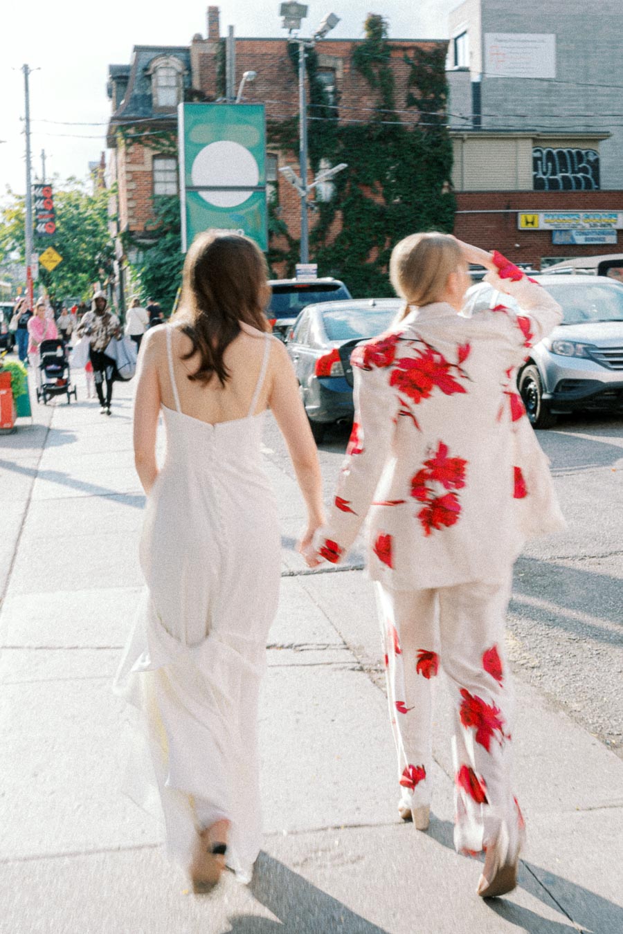 Two women dressed in elegant outfits, holding hands and walking down a sunlit urban street. One in a white dress, the other in a floral-patterned suit.