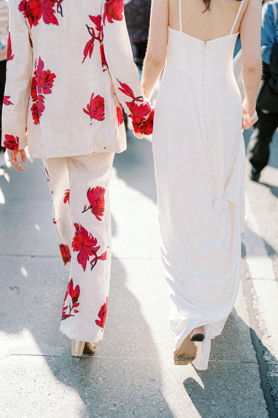 A couple walking hand-in-hand down a sunlit street, one wearing a floral print suit with red flowers and the other in an elegant white dress, showcasing fashion-forward wedding attire.