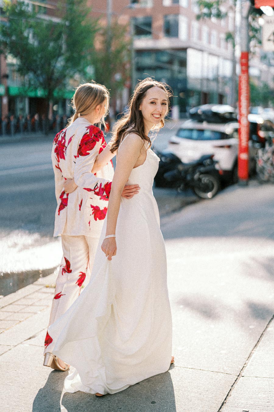 Two people walking outdoors, one wearing a white gown and the other in a floral suit, on a sunny day in an urban setting.