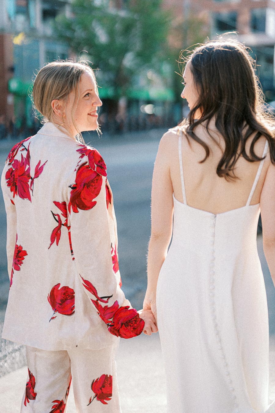 Two women holding hands, one in a floral suit and the other in a white dress, walking outdoors on a sunny day.