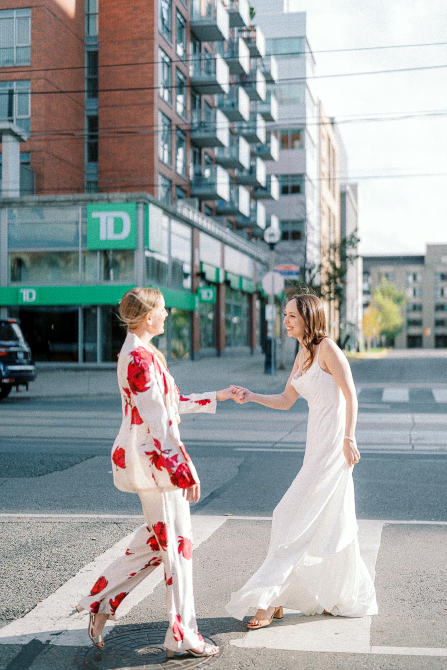 Two women greeting each other on a city street, one in a floral suit and the other in a white dress, in front of a modern urban building.