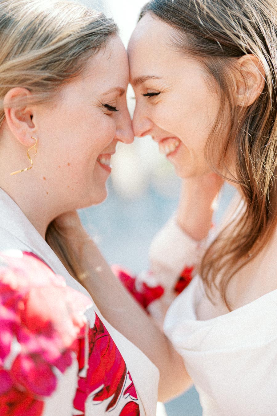 Two happy women touching foreheads, smiling in intimate and joyful moment, wearing white and floral outfits, captured in soft, natural lighting.