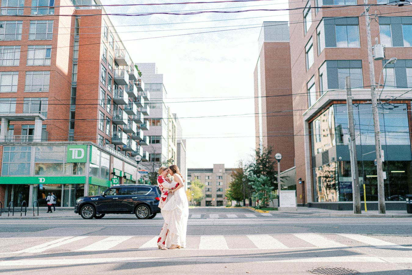 A couple embracing in wedding attire on a city street crosswalk, surrounded by modern buildings and a parked SUV, with a TD bank visible in the background.