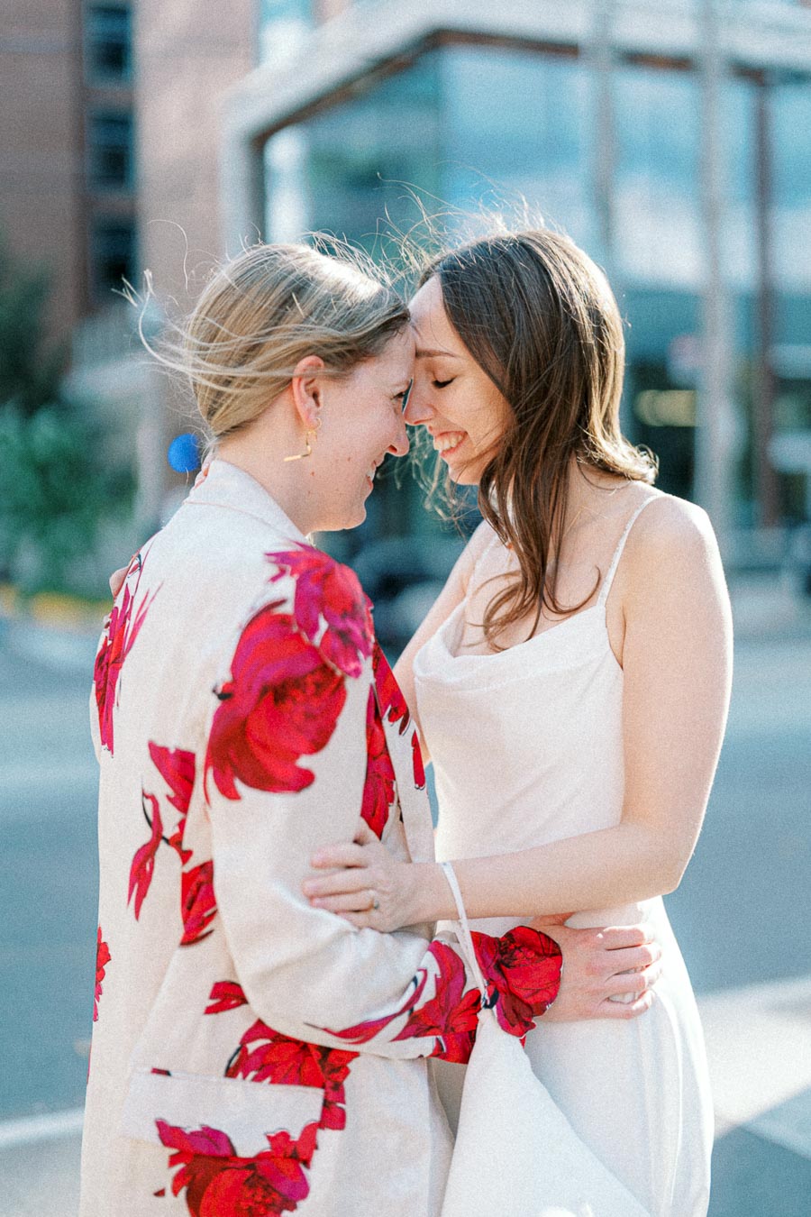 A loving couple embraces and smiles while leaning in for a tender moment on a sunny day in an urban setting. One wears a white dress, and the other has a floral-patterned jacket.