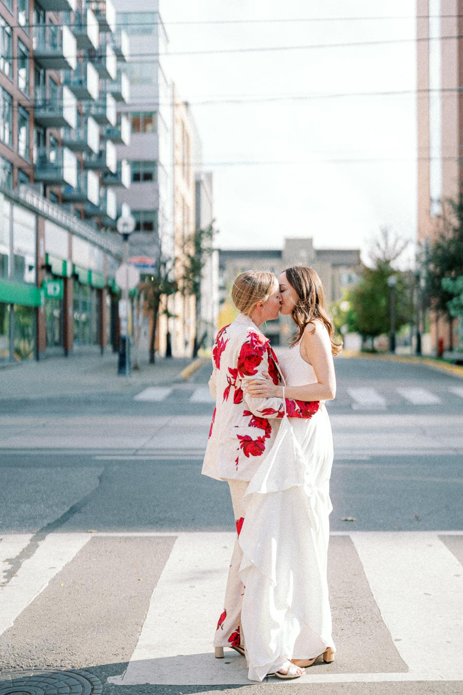 Couple sharing a kiss on a city street, featuring one in a floral suit jacket and another in a flowing white dress, symbolizing love and diversity in urban settings.