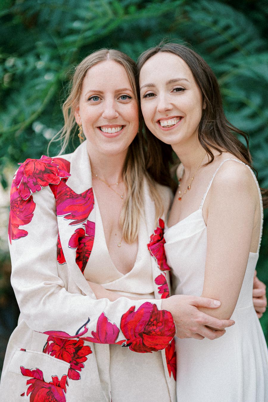 Two smiling women embracing in a garden setting, one wearing a floral-patterned blazer, the other in a white dress.